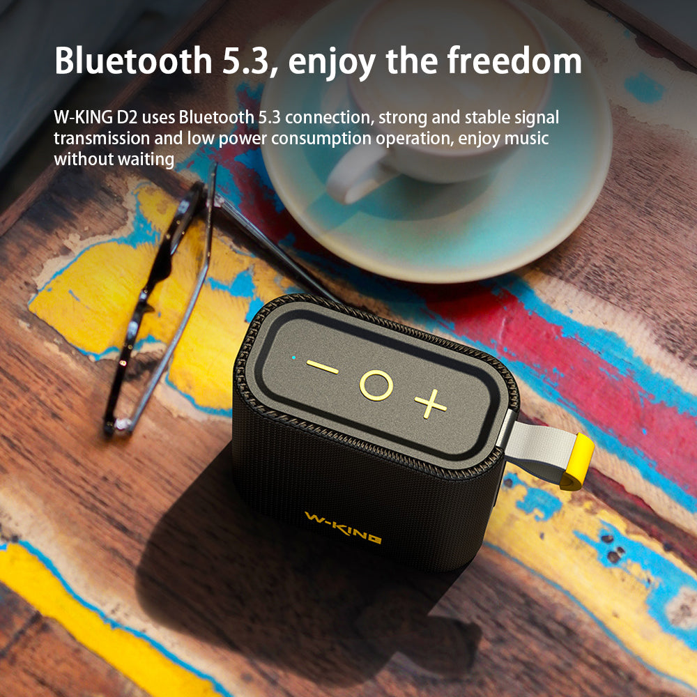 A compact and portable HeadphoneHeat W-King D2 Bluetooth Speaker With Wireless Connection in black rests on a vibrant wooden surface near a smartphone and coffee cup. Text highlights its Bluetooth 5.3 wireless connectivity for a strong, stable signal transmission.