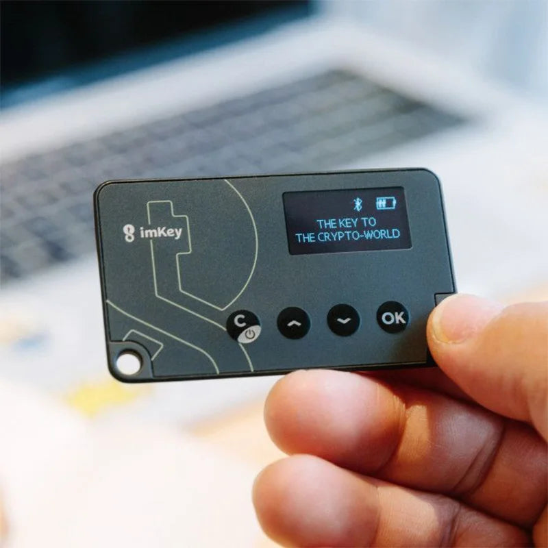 A person holds an ImKey Pro Crypto Hardware Wallet by the corner, a black electronic device with a small screen displaying "THE KEY TO THE CRYPTO-WORLD." The wallet features several buttons labeled C, arrow keys, and OK. In the background, a PVC wallet is visible next to a blurred laptop. Brand: HeadphoneHeat.