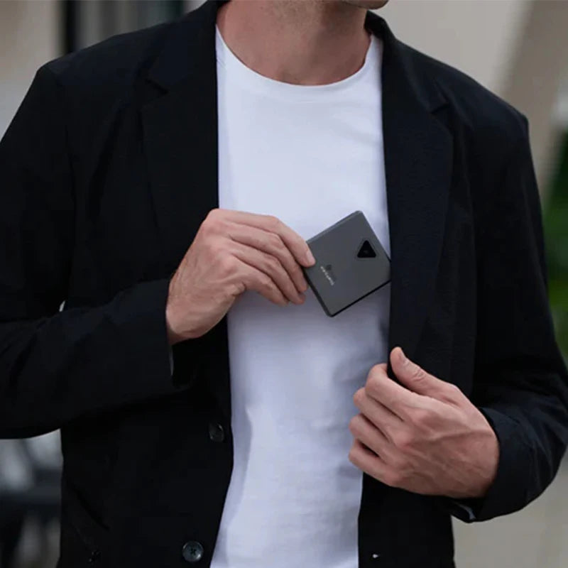 A person wearing a black blazer over a white shirt is placing an ELLIPAL Titan Mini security Cold Hardware Crypto Wallet, featuring the HeadphoneHeat brand logo and a black triangular button, into the inside pocket of their blazer. The background is blurred, focusing on the person's torso and hands.