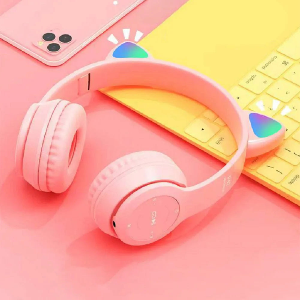 The Bluetooth VODOOL Wireless Cat Headphones with Ear Glow by HeadphoneHeat, in pink, rest on a yellow keyboard alongside a pink smartphone, all placed against a coordinating pink backdrop. These over-the-ear headphones feature vibrant lighting effects and provide dynamic sound quality.