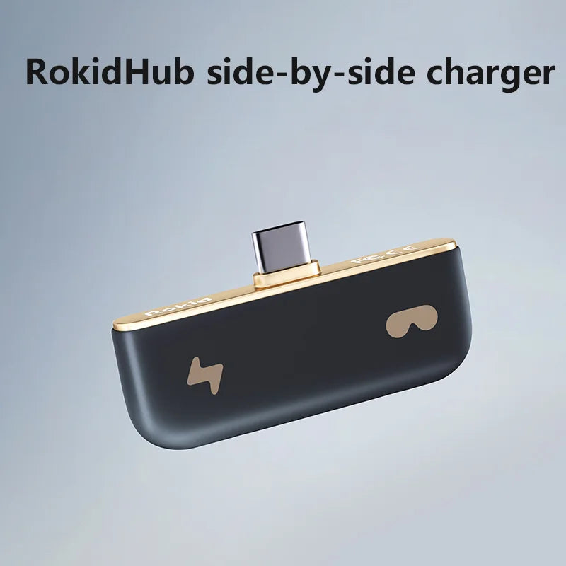 A product photo of the RokidHub side-by-side charger by HeadphoneHeat. The compact, black device, designed for Smart AR Glasses like the Rokid Air Smart VR Glasses, has a gold upper edge with a centrally positioned USB-C connector. It features a power symbol on the left and a VR headset symbol on the right. The background is a gradient of blue tones.