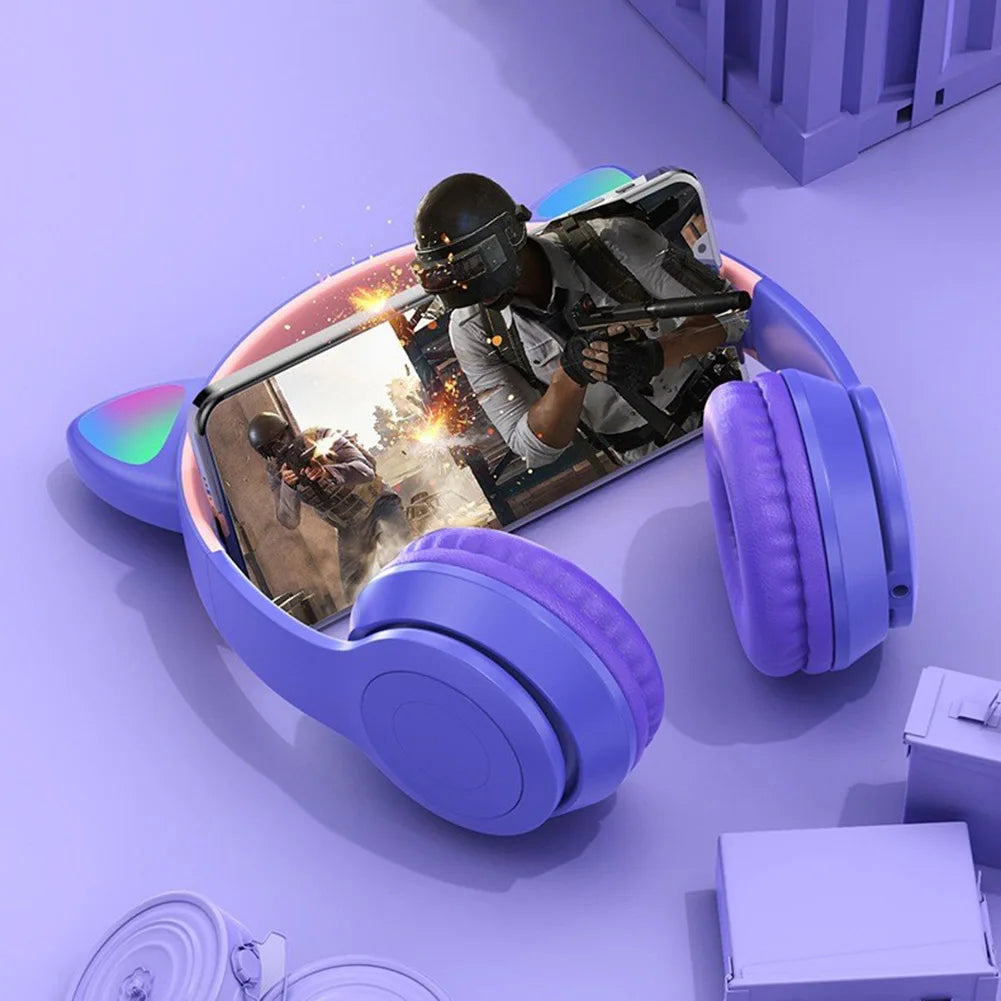 A smartphone showing a first-person shooter game is nestled within a pair of stylish HeadphoneHeat Bluetooth VODOOL Wireless Cat Headphones with Ear Glow. The game scene features a character in combat gear firing shots against a vivid purple backdrop, promising dynamic sound quality amidst the blurred surroundings.