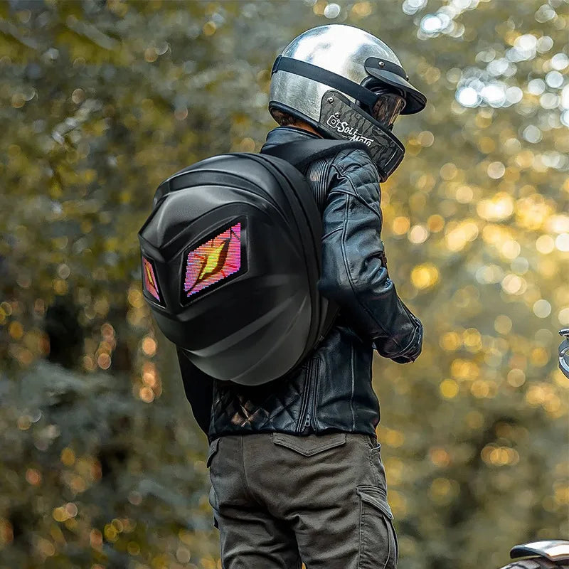 A person radiating an adventurous moto & biker style is walking outdoors, wearing a leather jacket and helmet, and carrying the HeadphoneHeat LED Turtle Shell Backpack. The lush greenery in the softly blurred background enhances the adventure-ready aura of this ABS material backpack.