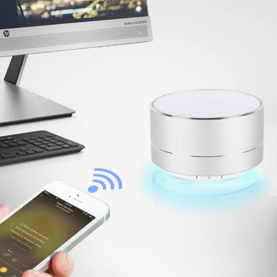 A desk setup includes a computer monitor, keyboard, and the Portable TOPK A10 Bluetooth Speaker from HeadphoneHeat.Com, boasting a durable design and emitting a soft blue light. A hand holds a smartphone playing music, wirelessly connecting to the speaker for superior sound quality.